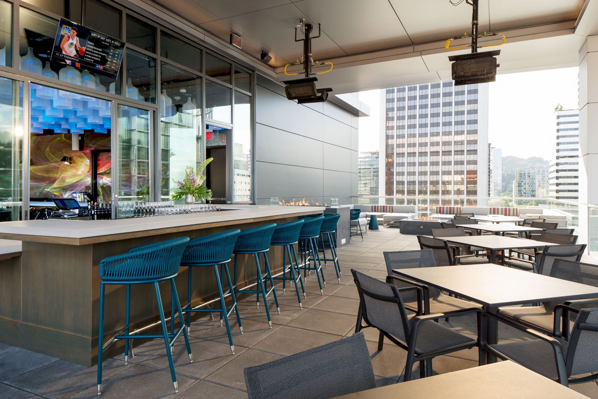 This image shows a modern rooftop bar with bar stools, tables, and chairs. It overlooks city buildings on a bright day.