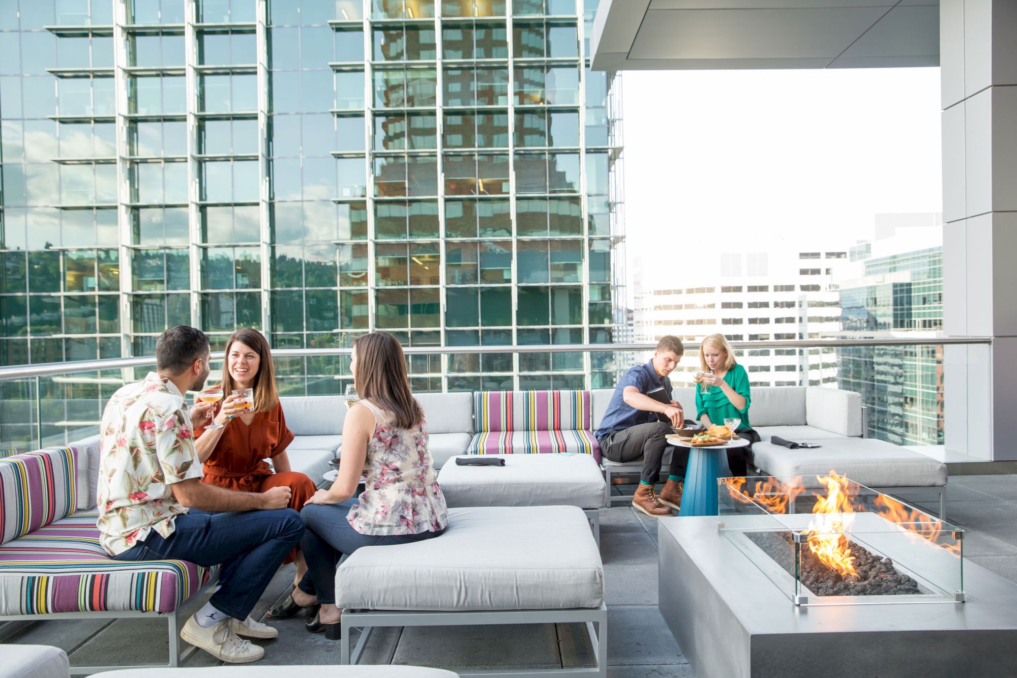 People sitting on a rooftop patio, conversing around modern fire pits, with city buildings in the background.