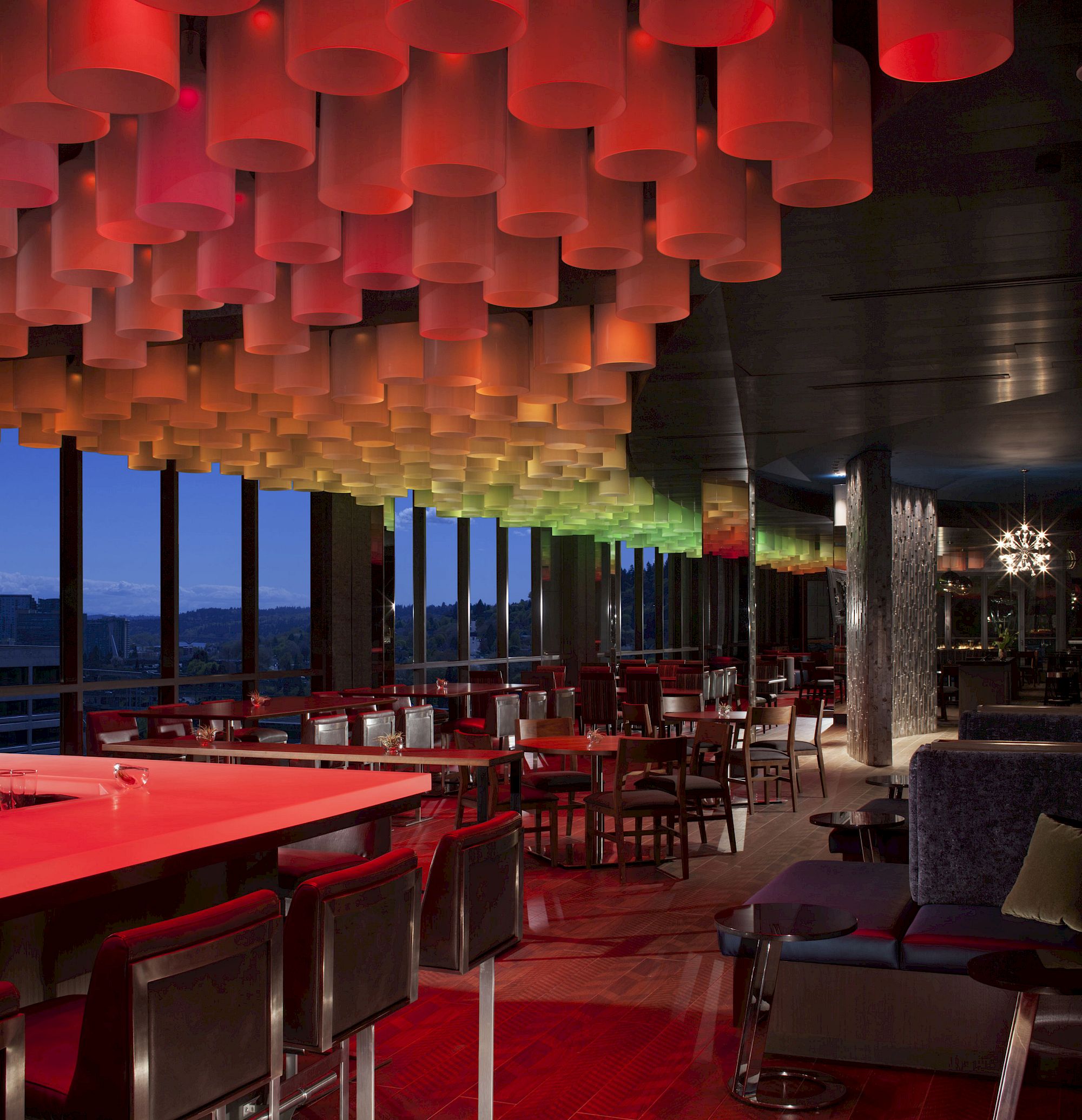 The image shows a modern, stylish bar or restaurant interior with colorful ceiling lights, high tables, and chairs, and large windows with a nighttime view.