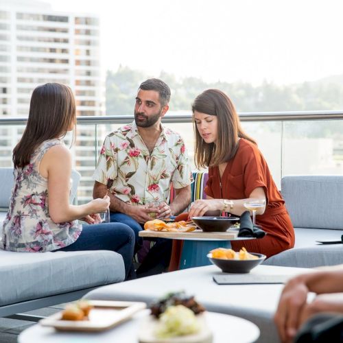 A group of people are sitting on a rooftop lounge, having a conversation and enjoying food, while one person sits separately and looks on.