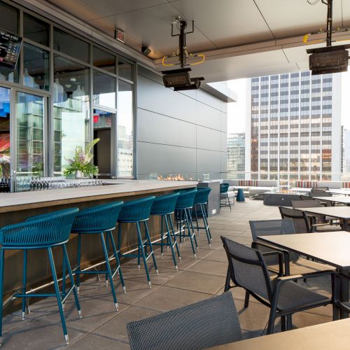 An outdoor rooftop bar with a long counter, blue bar stools, and multiple tables with chairs, set against a backdrop of skyscrapers.