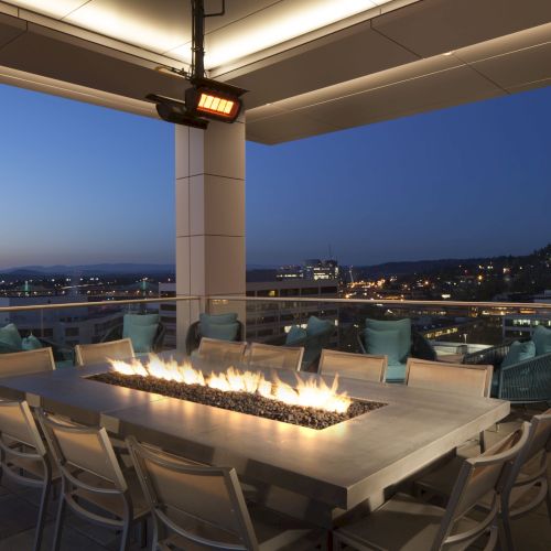 A rooftop patio at dusk featuring a long table with a built-in fire pit, surrounded by chairs, offering a view of the cityscape and distant mountains.