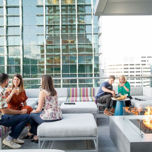 People are sitting on a rooftop patio with modern seating and a fire pit, surrounded by city skyscrapers.