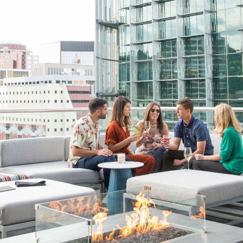 A group of five people sits on outdoor patio furniture, chatting and holding drinks. They are near a fire pit, with a cityscape as the background.