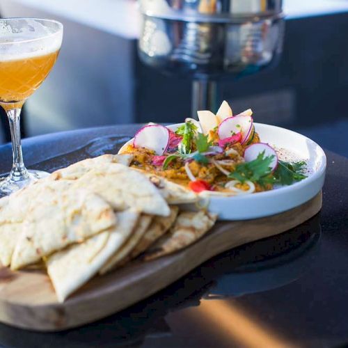 A platter with sliced flatbread and a colorful salad, paired with a cocktail, set on a round table outdoors.