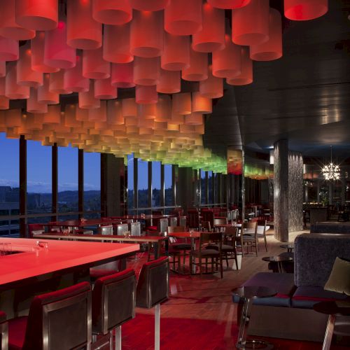 A trendy restaurant or bar with vibrant red and green hanging lights, sleek furniture, and large windows overlooking a nighttime cityscape.