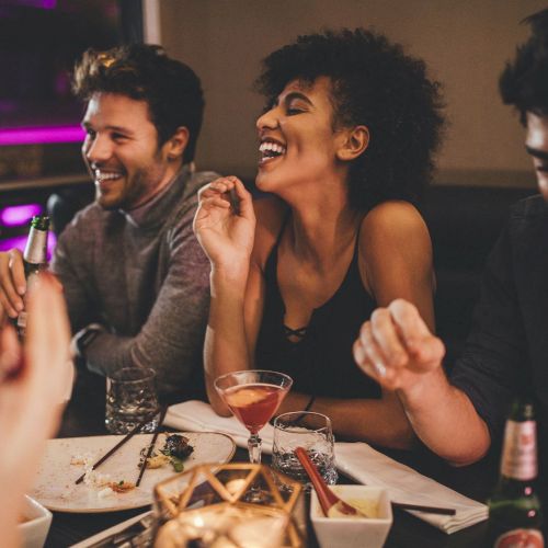 A group of friends is enjoying a night out together, laughing and having drinks at a dimly lit restaurant or bar, creating a lively atmosphere.