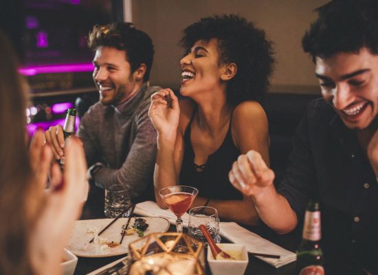 A group of friends is enjoying a night out together, laughing and having drinks at a dimly lit restaurant or bar, creating a lively atmosphere.
