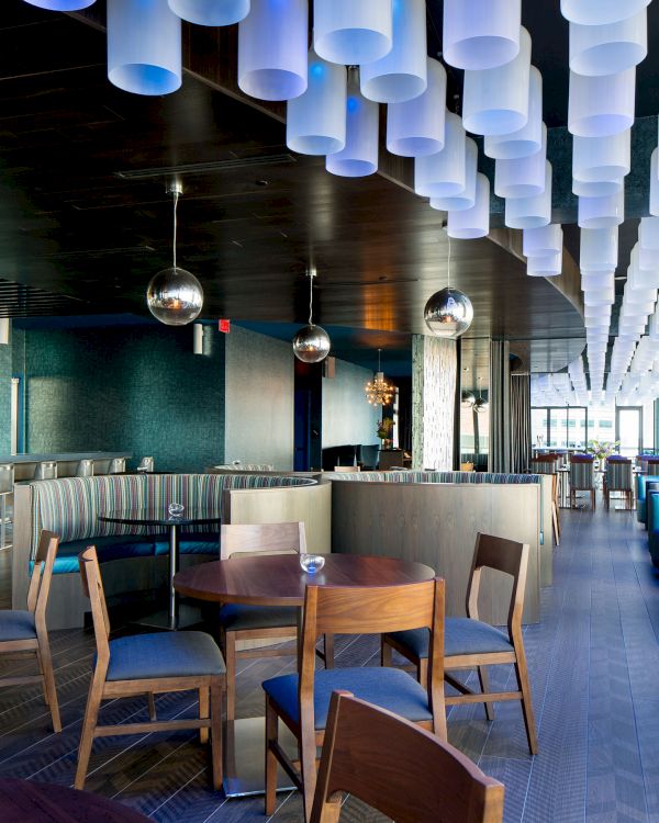 A modern, stylish lounge with wooden tables, blue seating, hanging globe lights, and cylindrical ceiling fixtures, featuring a bar area on the left.